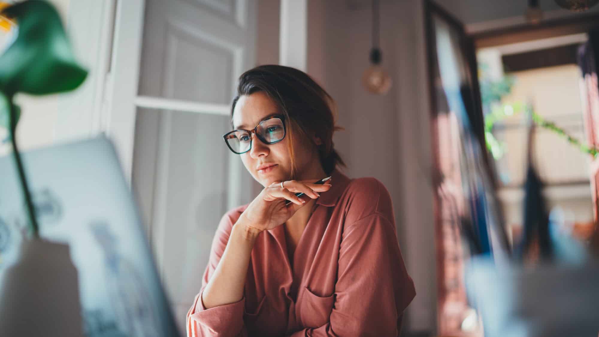 Woman with glasses, holding a pen and looking at laptop