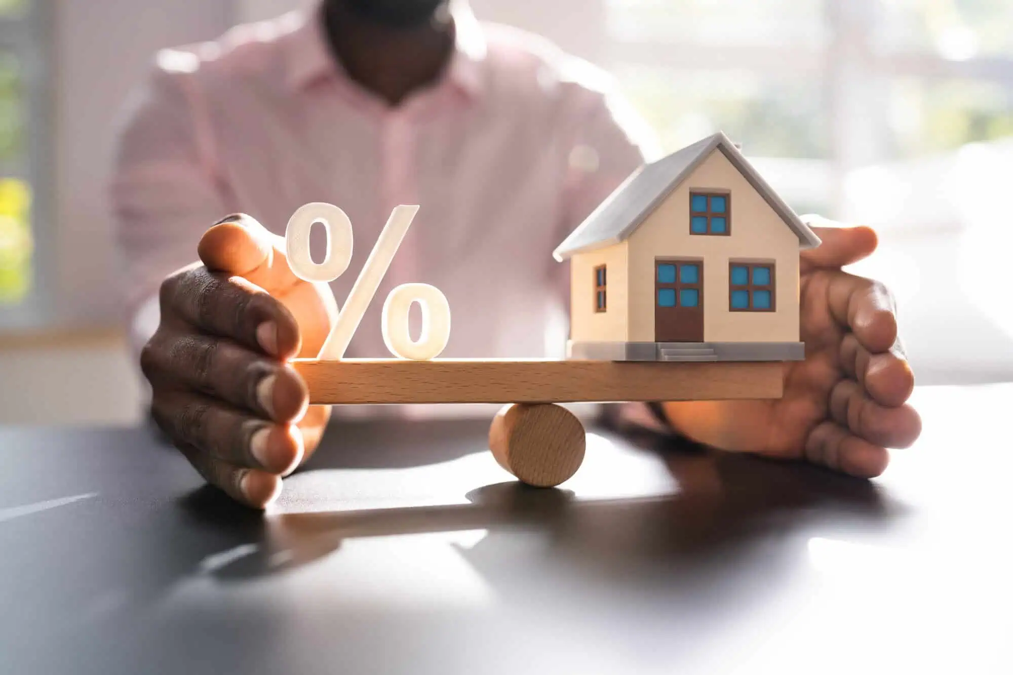 Person with model house and percent symbol on a piece of wood, balanced on a rounded piece of wood